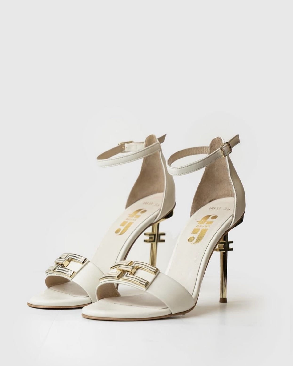 A pair of white leather heeled sandals with a gold buckle strap and the Feruza logo on the insole