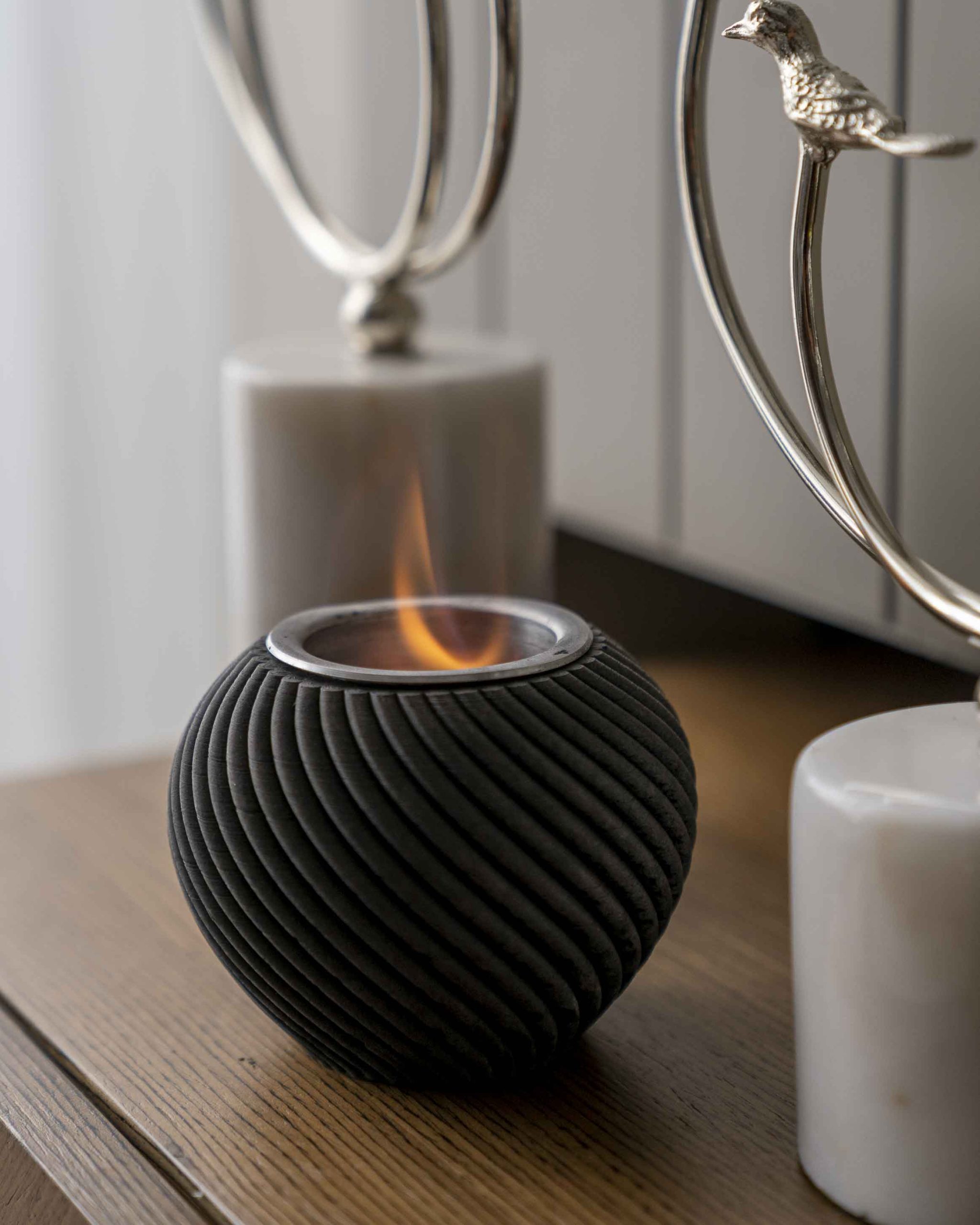 Spiral dokulu siyah ateş kabı ahşap konsolda yanıyor; arkada gümüş dekoratif objeler ve beyaz mumlar